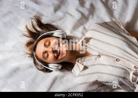 Crop Kind mit Kopfhörer einer Frau, die im Schlafzimmer auf dem Bett liegt und Musik hört, während die Augen geschlossen sind Stockfoto