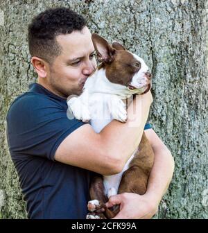 Ein junger hispanischer Mann umarmt und küsst einen entzückenden brindle Boston Terrier Welpe in der Nähe eines großen Baumstammes auf einem Sonniger Tag Stockfoto