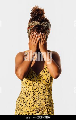 Lächelnde afroamerikanische Frau trägt Sommerkleid und trendige Stirnband Stehen in der Nähe von Wand in der Stadt und Bedeckung Gesicht beim Blick Bei der Kamera Stockfoto