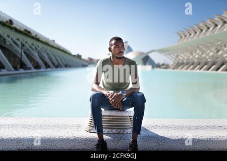 Nachdenkliche afroamerikanische Männer in lässiger Kleidung entspannend in der Nähe von Brunnen In der Stadt an sonnigen Tag und Blick weg Stockfoto