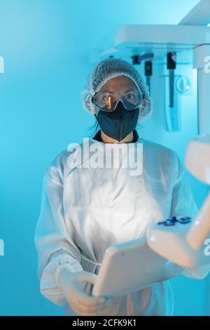 Professionelle Ärztin in medizinischer Uniform und Schutzmaske mit Brillen, die die Kamera betrachten, während sie moderne Diagnosegeräte in verwenden Krankenhaus Stockfoto