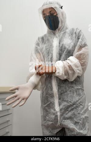 Weibliche medizinische Fachärztin in Schutzanzug mit Gesichtsschutz und Atemschutzmaske, die während der Vorbereitung auf die Arbeit Latexhandschuhe anziehen Coronavirus-Pandemie Stockfoto