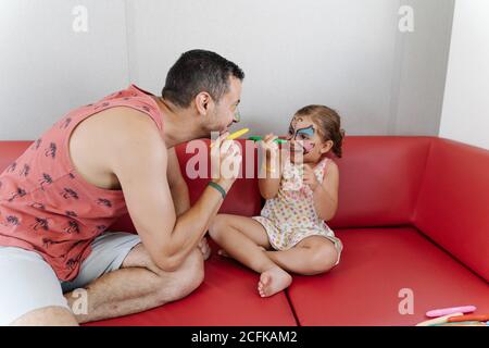 Seitenansicht von liebenswert Mädchen Malerei Gesicht von ruhigen Vater Beim gemeinsamen Entspannen auf dem Sofa und Spaß am Wochenende Stockfoto