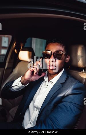Junge afroamerikanische männliche Unternehmer in eleganten formellen Anzug reden Auf dem Handy, während Sie mit dem Auto auf der Stadtstraße fahren Stockfoto