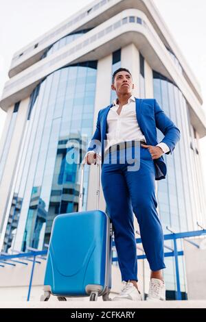 Von unten positive junge afroamerikanische männliche Unternehmer in Eleganter Anzug in der Nähe des Koffers auf der modernen Stadtstraße Stockfoto
