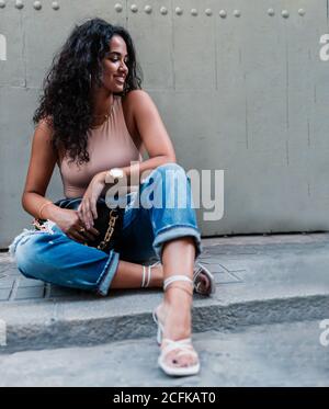 Voller Körper lächelnd junge lockig behaarte ethnische Brünette in Stylische zerrissene Jeans und mit Accessoires auf Steintreppen In der Nähe der grauen Wand Stockfoto