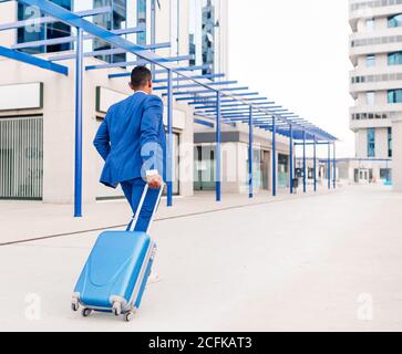 Ganzkörper-Rückansicht von nicht erkennbaren ethnischen Geschäftsmann in elegant Formell Anzug ziehen Koffer beim Gehen Stockfoto