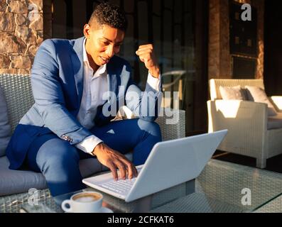 Fröhliche junge afroamerikanische Geschäftsmann in formelle Kleidung mit Laptop Und gestikulieren mit Faust bis glücklich mit großen Nachrichten während Sitzen mit einer Tasse Kaffee in der Lounge Zone der modernen Cafeteria Stockfoto