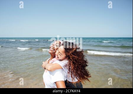 Entzückt schwarz Mutter Huckepack junge Tochter beim mit Spaß mit Geschlossene Augen am Meer im Sommer Stockfoto