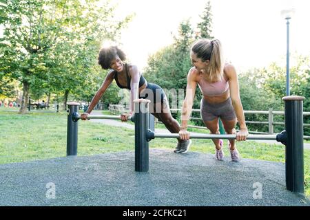 Ganzkörper positive junge multirassische weibliche Freunde in Sportswear tun Push-up-Übung auf Metallstangen während Fitness-Training in Sommerpark Stockfoto