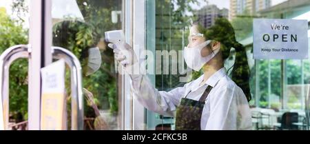 Asiatische Kellnerin mit Gesichtsmaske, die vor dem Besuch im Restaurant Temperatur auf den Kunden nimmt. Neues normales Restaurant Lifestyle Konzept nach cor Stockfoto