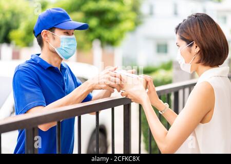 Asian liefern Mann in blauem Hemd Handhabung Paket und geben an junge Frau Kunden zu Hause. Sie tragen Gesichtsmaske, um Coronavirus COVID-19 spreadi zu reduzieren Stockfoto