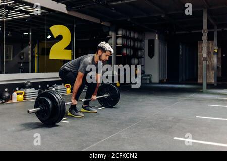 Ganzkörper-Seitenansicht des konzentrierten jungen bärtigen männlichen Athleten Vorbereitung für das Heben schwere Langhantel während intensives Training in zeitgenössischen Sportklub Stockfoto