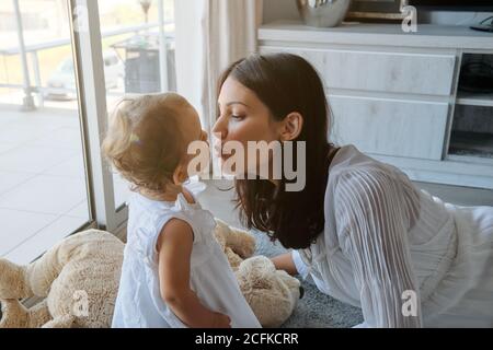 Frau liegt auf dem Boden und küsst ein kleines Mädchen hinein Ein weißes Kleid im Esszimmer des Hauses Stockfoto