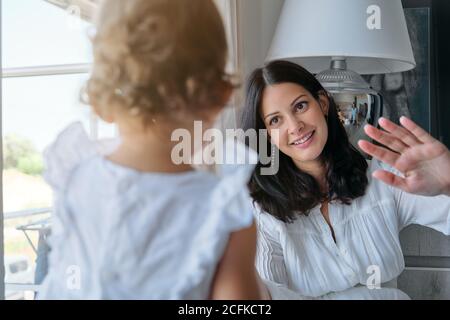 Mutter winkt einem kleinen Mädchen in einem weißen Kleid zu Sitzen auf dem Boden eines Esszimmers in einem Haus Stockfoto