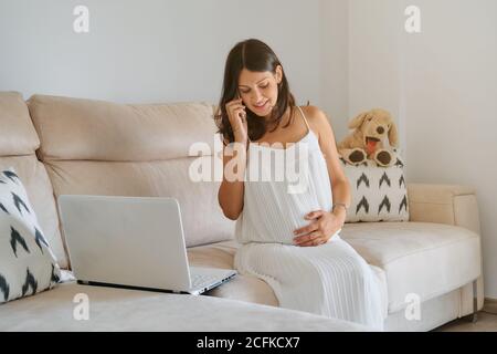 Eine Schwangere berührt ihren Bauch mit der Hand, während sie am Telefon vor einem Laptop auf einer Couch in einem Haus sitzt Stockfoto