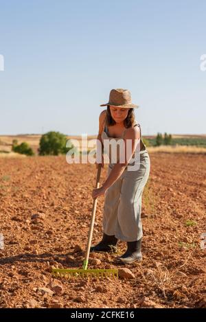 Bäuerin in Sonnenhut mit Rechen auf dem Boden des Feldes Während der Arbeit im Dorf im Sommer Stockfoto