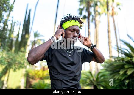 Junger afroamerikanischer Athlet in Sportkleidung, der Musik hört Mit seinen Kopfhörern halten die Hände auf den Kopfhörern und geschlossen Augen im grünen tropischen Park stehen Stockfoto