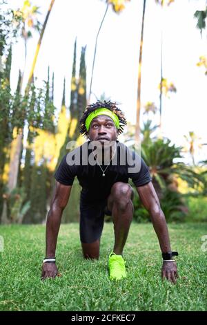 Entschlossener junger afroamerikanischer Athlet im stylischen Sportswear Stehen In niedriger Startposition auf grünem Rasen während der Vorbereitung Laufen im Park Stockfoto