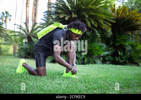 Ganzkörper-Seitenansicht des jungen afroamerikanischen Sportlers in Stylische Sportbekleidung mit hellem Rucksack und Stirnband, das Schnürsenkel bindet Sneaker während der Vorbereitung auf das Training im grünen tropischen Park Stockfoto