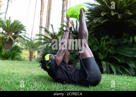 Junger afroamerikanischer Sportler in stylischer Activewear auf Grün sitzend Gras und Stretching-Übungen nach dem Laufen im Park Stockfoto