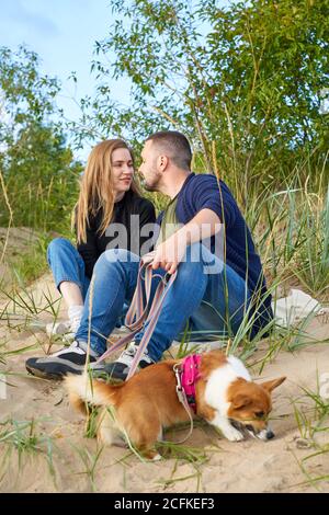 Junge glückliche Paar von Mann und Frau mit Corgi Hund sitzen am Sand. Männlich küssen schön weiblich. Zwei Personen in legerer Kleidung genießen schönen sonnigen Nachmittag am Strand Stockfoto
