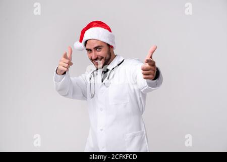 Jungen gutaussehenden Arzt in Weiß uniforme und Santa Claus hat im Studio auf weißem Hintergrund Lächeln und zeigt mit dem Finger in die Kamera Student der Medizin Stockfoto