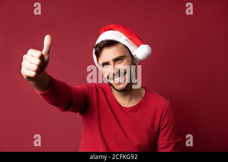 Junge stattlichen kaukasischen Kerl im roten Pullover und Nikolausmützen steht auf roten Hintergrund im Studio und zeigt den Daumen nach oben. Close up Portrait europäischen Kerl wi Stockfoto