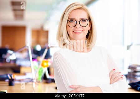Porträtaufnahme einer attraktiven Geschäftsfrau, die mit im Büro gekreuzten Armen steht, während sie die Kamera betrachtet und lächelt. Stockfoto