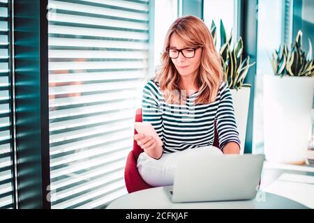 Porträtaufnahme einer selbstbewussten Geschäftsfrau mit Textnachrichten, während sie auf einem Stuhl sitzt und von zu Hause aus arbeitet. Heimbüro. Stockfoto