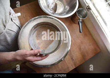 Eine Köchin in Schürze siebt Mehl in einer Metallschüssel. Vorbereitung für die Herstellung eines Teigs zum Backen. Hausgemachte Patisserie. Hochwertige Fotos Stockfoto