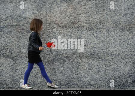 Kleine Stadt stilvolle Mädchen in lässigen Herbst Kleidung zu Fuß entlang graue Granitwand mit rotem Wind Spinner. Leerzeichen für Text. Stockfoto