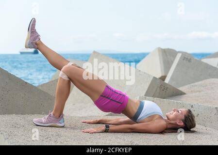 Seitenansicht der athletischen Frau mit starkem Körper, der Brücke tut Trainieren Sie im Sommer mit Gummizug am Wellenbrecher Stockfoto