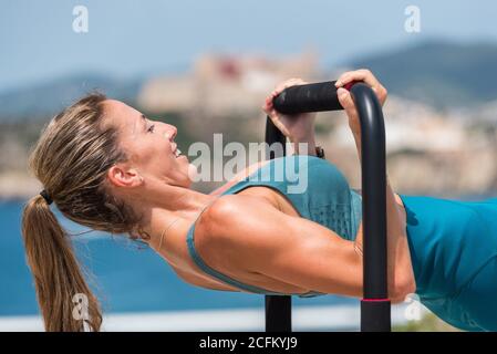 Seitenansicht der starken Frau in Sportswear tun Pull-ups Während des Trainings an sonnigen Tagen an der Küste Stockfoto