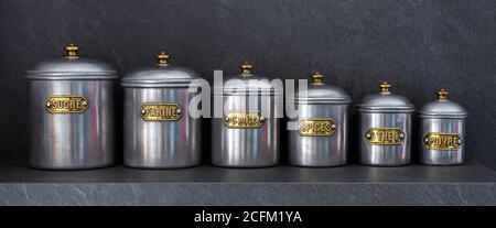 Set aus sechs Zinngläsern in verschiedenen Größen, aufgereiht auf einem Regal mit den Aufschriften 'SUGAR', 'MEHL', 'KAFFEE', 'SPICES', 'TEE' und 'PFEFFER'. Stockfoto