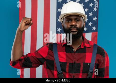 Junger Baumeister oder Vorarbeiter, der seinen starken Arm eingebogen zeigt Ellenbogen gegen US-Flagge Stockfoto