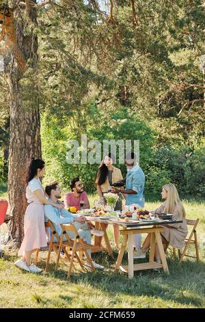 Erholsame junge Freunde sitzen und stehen am Tisch unter serviert Kiefernbaum Stockfoto