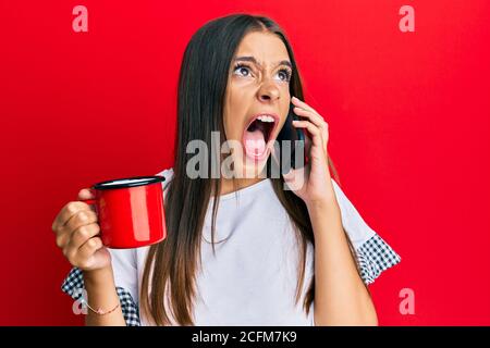 Junge hispanische Frau im Gespräch auf dem Smartphone und trinken eine Tasse Kaffee wütend und verrückt schreien frustriert und wütend, schreien mit Wut schaut Stockfoto