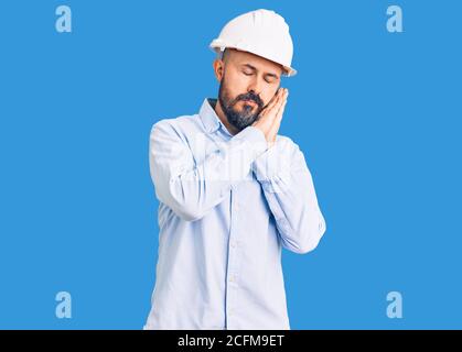 Junger, hübscher Mann, der Architekt und Hardhut trägt und müde schläft und mit Händen zusammen posiert, während er mit geschlossenen Augen lächelt. Stockfoto