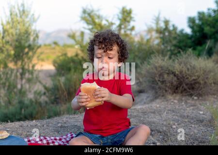 Lustige Picknick-Situation eines Jungen, der ein Sandwich isst Die Berge mit Begeisterung sitzen auf einem rot-weiß Karierte Tischdecke Stockfoto
