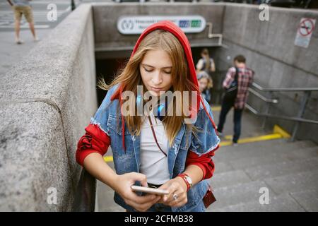 Eine junge schöne Frau in rotem Hoodie steht am Eingang der U-Bahn mit Handy in den Händen. Stockfoto