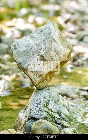 Ausgewogener Felsen am Bach. Ein würfelförmiges Gestein wird natürlich auf einem Felsen in einem Bach, der von Wasser umgeben ist, ausgeglichen. Klassische Steinbalance. Stockfoto