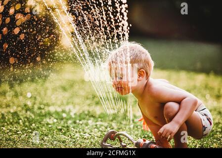 Kleiner Junge, der die Zunge herausstreckt und Wasser aus dem Sprinkler herausstreckt Garten Stockfoto