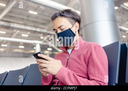 Ein Typ wartet in der Flughafenlounge in einer medizinischen Maske auf einen Flug. Flugreisen während der Coronavirus-Pandemie. Überprüfen seines Telefons und Stockfoto