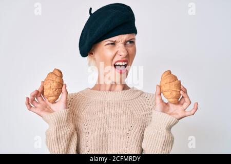 Junge kaukasische Frau im französischen Look mit Baskenmütze hält Croissants wütend und verrückt schreien frustriert und wütend, schreien vor Wut. Wut und Stockfoto