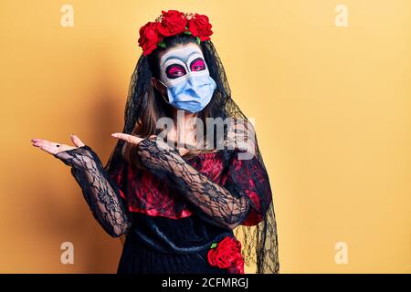 Junge Frau trägt Tag der Toten Kostüm trägt medizinische Maske erstaunt und lächelt an der Kamera, während mit der Hand und zeigt mit dem Finger. Stockfoto
