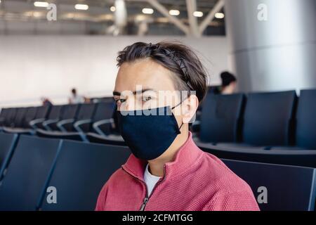 Ein Typ wartet in der Flughafenlounge in einer medizinischen Maske auf einen Flug. Flugreisen während der Coronavirus-Pandemie. Überprüfen seines Telefons und Stockfoto