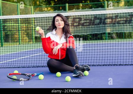 Junge hübsche Frau, die auf dem Tennisnetz sitzt und sich nach dem Training entspannt. Porträt der Sportlerin auf dem blauen Tennisplatz. Gesundes Leben. Stockfoto