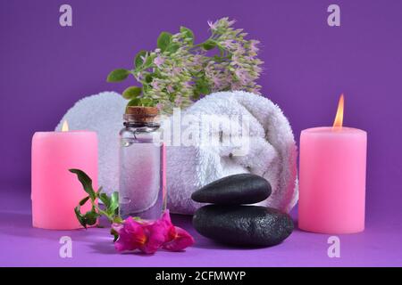 Rosa Kerze brennend, Handtuch mit Blumen, kleine Flasche mit Balsam, und schwarze Steine auf einem lila Hintergrund. Spa- und Wellness-Konzept Stockfoto