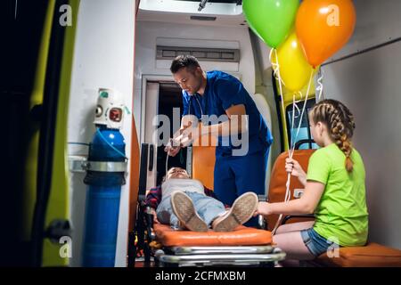 Bewusstlose Mutter auf einer Bahre in einem Krankenwagen liegend, gibt ihr ein Sanitäter eine Sauerstoffmaske und spricht mit ihrer kleinen Tochter, um sie zu beruhigen. Stockfoto